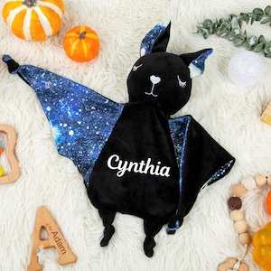 May include: A black plush bat security blanket featuring a galaxy print on the inner wings and ears. The name Cynthia is embroidered in white script on the center of the black body. The bat has closed eyes and a white heart-shaped nose.