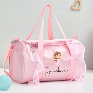 May include: A light pink cylindrical duffel bag featuring a graphic of a ballerina in a pink tutu and the name Jackie printed in black script. The bag has two top handles, a detachable shoulder strap, and decorative pink tulle ruffles.