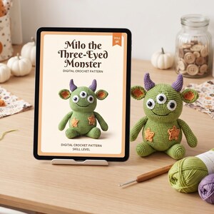 May include: A tablet displaying a digital crochet pattern titled Milo the Three-Eyed Monster, featuring a green crocheted monster with purple horns, three eyes, and orange star patches, alongside a physical version of the same crocheted monster.