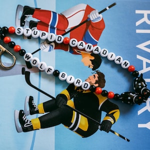 Op de afbeelding: Een kralen sleutelhanger met witte letterkralen die STUPID CANADIAN WOLF BIRD spellen, afgewisseld met kleine zwarte en rode ronde kralen. De sleutelhanger bevat een zilverkleurige metalen karabijnhaak en een zwart-witte dierenbedel.