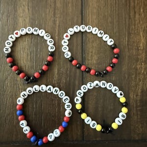 Op de afbeelding: Vier handgemaakte kralenarmbanden met witte letterkralen die MOY POMIDOR, MOY LYUBIMIY, HOLLANDER 24 en ROZANOV 81 spellen. De armbanden bevatten diverse gekleurde accentkralen in rood, zwart, blauw en geel, samen met kleine hart- en stersymbolen.