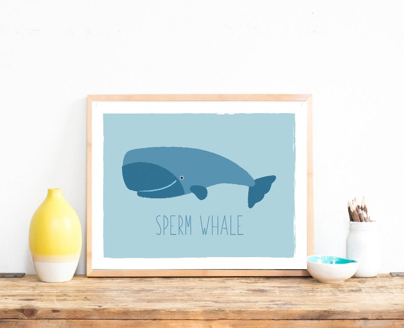 May include: A framed print featuring a blue sperm whale illustration on a light blue background. The text "SPERM WHALE" is printed below the whale. The artwork is displayed on a wooden surface.