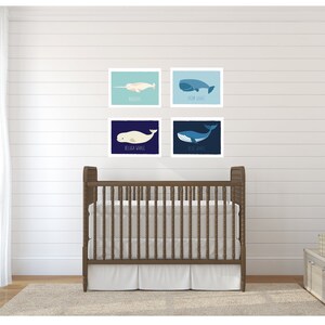 May include: Four framed whale art prints in a nursery setting. The prints feature illustrations of different whales, including a narwhal and a blue whale, in shades of blue and white. A wooden crib and neutral decor complete the scene.