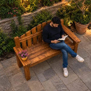 Puede incluir: Un banco de madera con respaldo de listones y un reposabrazos plano que presenta una pequeña maceta con flores rosas. El banco está construido con tablones de madera teñida y ofrece asiento para una persona que lee un libro con pantalones vaqueros.