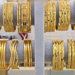 May include: A collection of gold bangles displayed on two horizontal gray velvet bars. The bangles feature various textures, including smooth, wavy, and intricate geometric patterns, all crafted in a polished yellow gold finish.