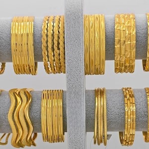 May include: A collection of twelve sets of gold-colored bangles displayed on a gray velvet stand. The bangles feature various textures including twisted, wavy, and geometric patterns, with some sets showing smooth surfaces and others displaying intricate engravings.