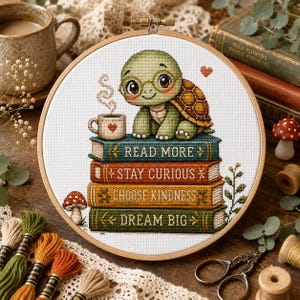 May include: A circular wooden embroidery hoop containing a cross-stitch design of a small green turtle wearing round glasses, sitting atop a stack of four colorful books. The books feature the text READ MORE, STAY CURIOUS, CHOOSE KINDNESS, and DREAM BIG.