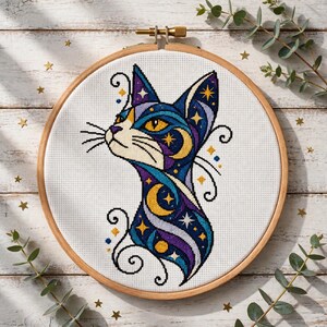 Puede incluir: Un bordado de punto de cruz con el perfil de un gato en un bastidor de madera. El gato presenta un patrón celestial en tonos azul profundo, morado y amarillo, incorporando lunas crecientes y estrellas con líneas negras curvas.