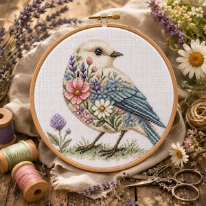 May include: A circular wooden embroidery hoop containing a cross-stitch pattern of a bird with cream-colored feathers, decorated with blue, pink, and white floral motifs. The bird stands on a small patch of green grass with a purple clover flower.