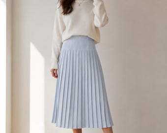 Puede incluir: Un suéter de punto color crema de manga larga combinado con una falda plisada midi de color azul claro y zapatillas blancas. El suéter presenta un cuello redondo y puños acanalados, mientras que la falda tiene cintura alta y pliegues verticales.