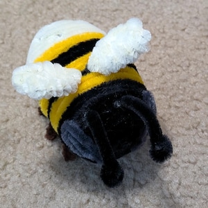 May include: A small bee figurine crafted from fuzzy pipe cleaners. The body features alternating horizontal stripes of bright yellow and black. It has two white, textured wings extending from the sides and two black antennae protruding from the front.
