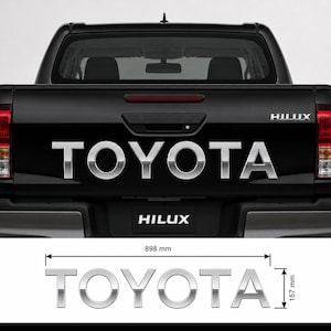 Peut inclure: Un ensemble d'emblèmes TOYOTA chromés pour hayon de pick-up. Les lettres sont présentées sur le hayon d'un véhicule noir et séparément avec des lignes de cote indiquant une largeur de 898 mm et une hauteur de 157 mm pour l'ensemble du lettrage.