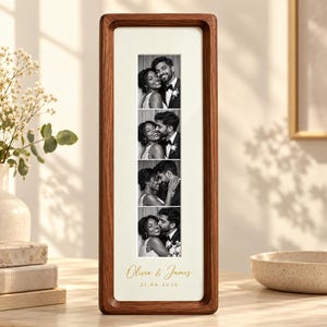Op de afbeelding: Een verticale rechthoekige houten fotolijst met een donkerbruine afwerking die een strook van vier zwart-witfoto's van een koppel bevat. Onder de foto's staan de tekst Olivia & James en de datum 21.06.2026 in goudkleurig handschrift gedrukt.