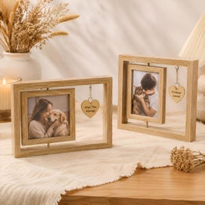 May include: Two light-colored wooden rotating picture frames. The left frame holds a photo of a person with a dog and a heart charm reading "Miss You Always." The right frame holds a photo of a person with a cat and a heart charm reading "Forever Loved."