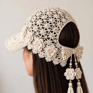 May include: A cream-colored crochet baseball cap featuring an intricate star-patterned knit, embellished with small gold beads and floral appliqués. The back includes a decorative bow with dangling beaded strands and long cream-colored tassels.