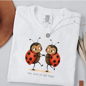 May include: A white t-shirt featuring a graphic of two cartoon ladybirds holding hands. The ladybirds have red wings with black spots and brown bodies. Below the illustration, the text reads "the last of the bugs" in a simple black font.