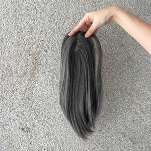 May include: A hair topper featuring straight, shoulder-length hair in a blend of dark gray and silver tones. The piece includes a monofilament base at the crown, designed to mimic a natural scalp appearance with a visible center part.