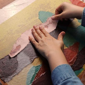 May include: A rectangular sheet of textured paper featuring a collage of overlapping, torn-edge sections in muted shades of yellow, brown, dark green, and red. A light pink, irregular strip of paper is being carefully pressed onto the center surface.