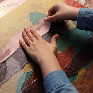 May include: A rectangular sheet of textured paper featuring a layered collage of torn paper pieces in shades of yellow, pink, dark brown, teal, and red. The surface has a fibrous, handmade appearance with visible overlapping edges and varied color blocks.
