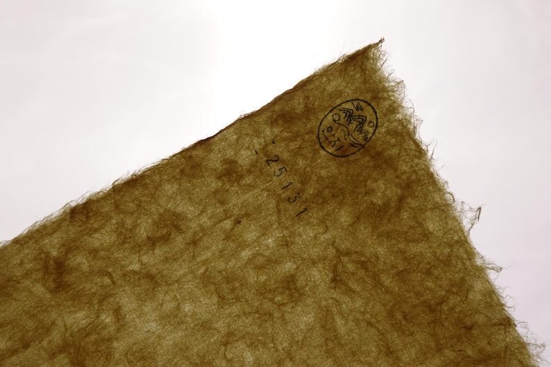 May include: A sheet of olive-green handmade paper with a fibrous, textured surface and deckled edges. A circular black ink stamp featuring a mountain illustration and the text 25131 is visible in the upper right corner of the paper.