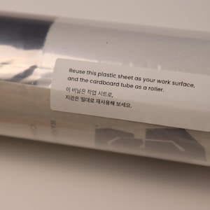 May include: A cylindrical cardboard tube wrapped in a clear plastic sheet. A white rectangular label is affixed to the surface with the text: Reuse this plastic sheet as your work surface, and the cardboard tube as a roller. 이 비닐은 작업 시트로, 지관은 밀대로 재사용해 보세요.