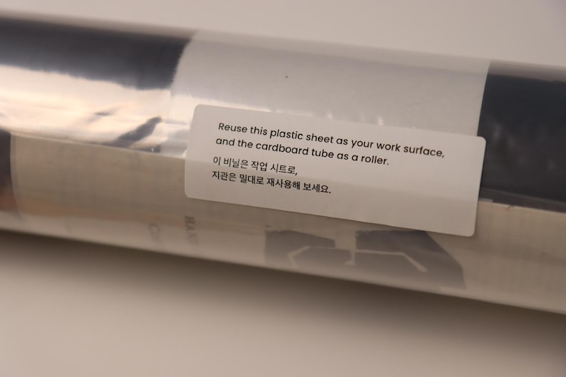 May include: A cylindrical cardboard tube wrapped in a clear plastic sheet. A white rectangular label is affixed to the surface with the text: Reuse this plastic sheet as your work surface, and the cardboard tube as a roller. 이 비닐은 작업 시트로, 지관은 밀대로 재사용해 보세요.