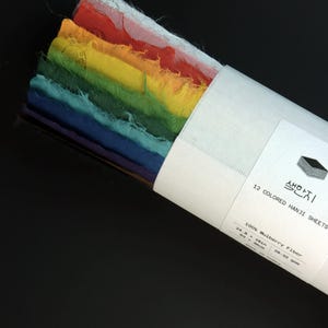 May include: A stack of 12 colored Hanji sheets made of 100% mulberry fiber, featuring raw, deckled edges in a rainbow gradient. The paper is secured with a white paper band labeled 12 COLORED HANJI SHEETS, 24.8 x 18 inches, 28-32 gsm.