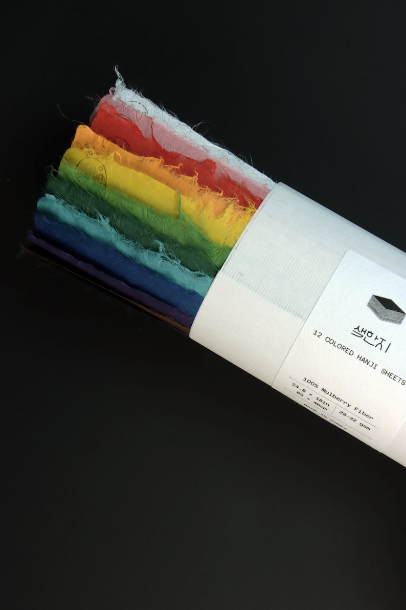 May include: A stack of 12 colored Hanji sheets made of 100% mulberry fiber, featuring raw, deckled edges in a rainbow gradient. The paper is secured with a white paper band labeled 12 COLORED HANJI SHEETS, 24.8 x 18 inches, 28-32 gsm.