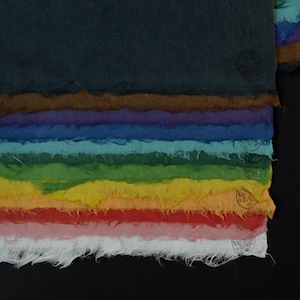May include: A stack of rectangular handmade paper sheets with rough, deckled edges, arranged in a gradient of colors including dark blue, brown, purple, blue, teal, green, yellow, orange, red, pink, and white, each featuring a small circular stamp.