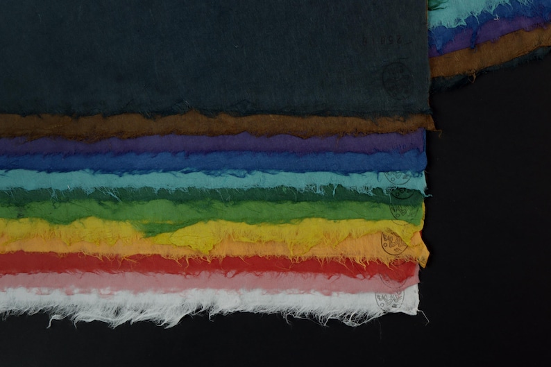 May include: A stack of rectangular handmade paper sheets with rough, deckled edges, arranged in a gradient of colors including dark blue, brown, purple, blue, teal, green, yellow, orange, red, pink, and white, each featuring a small circular stamp.