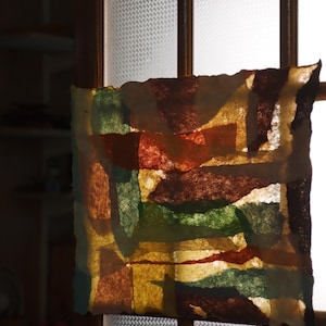 May include: A rectangular piece of translucent paper featuring an abstract collage of overlapping shapes in shades of deep brown, forest green, mustard yellow, and burnt orange, with rough, deckled edges, backlit to reveal the layered texture.