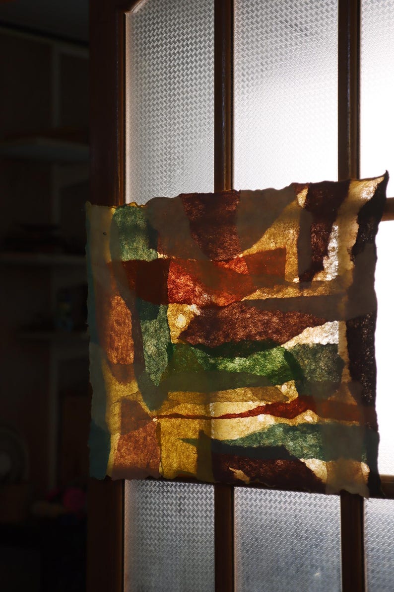 May include: A rectangular piece of translucent paper featuring an abstract collage of overlapping shapes in shades of deep brown, forest green, mustard yellow, and burnt orange, with rough, deckled edges, backlit to reveal the layered texture.