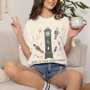 May include: A cream-colored t-shirt featuring a central dark green grandfather clock graphic surrounded by illustrations of a ballerina, a nutcracker, a mouse soldier, and a horn. The text "WHEN THE CLOCK STRIKES MIDNIGHT" is printed in dark green.