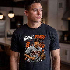 Puede incluir: Una camiseta negra de manga corta con un estampado gráfico de un jugador de béisbol con uniforme gris, el número 8, el texto GAME READY y el logotipo de los Baltimore Orioles. El diseño incluye detalles de salpicaduras de pintura naranja y blanca.