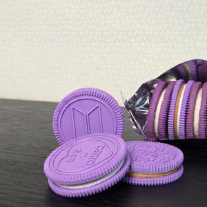Op de afbeelding: Een stapel paarse sandwichkoekjes met witte crèmevulling, voorzien van het BTS-logo en de tekst OREO in reliëf op het oppervlak. Eén koekje staat rechtop, terwijl andere horizontaal gestapeld liggen naast een open metalen folieverpakking.