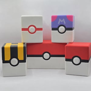 May include: A collection of five cube-shaped containers designed to resemble various spherical capture devices. The boxes feature white, red, yellow, black, and purple colour schemes with a central circular button and the letter M on one.