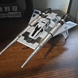 Peut inclure: Un modèle réduit de vaisseau spatial de type chasseur stellaire en plastique gris et noir, doté de deux longs canons pointus vers l'avant, d'un cockpit central et d'ailes angulaires. Le véhicule présente un design mécanique détaillé.