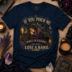 May include: A navy blue t-shirt featuring a graphic illustration of a person sleeping in bed with a black cat. The text reads: IF YOU PINCH ME WHILE I'M SLEEPING YOU WILL LOSE A HAND. The design includes a candle, books, and crescent moons.
