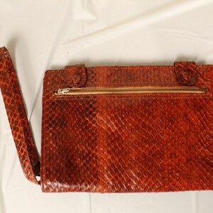 May include: A rectangular rust-colored clutch bag featuring a textured snakeskin pattern. The bag includes a matching wrist strap attached to the side and a horizontal gold-toned metal zipper closure across the upper portion of the front panel.