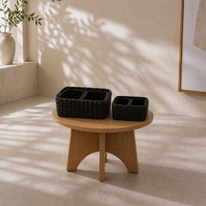 May include: Two rectangular storage baskets crafted from dark charcoal woven material, each featuring two internal compartments. The baskets are positioned on a light-toned wooden table with a circular top and four sturdy, arched support legs.