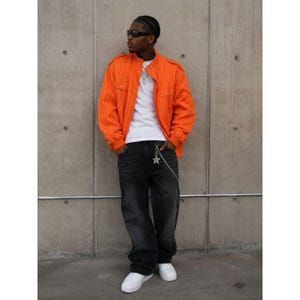 May include: A bright orange long-sleeve jacket with vertical stripes and shoulder epaulets worn over a plain white t-shirt. The outfit includes loose-fitting dark gray denim jeans, white sneakers, dark sunglasses, and a silver star-shaped chain.