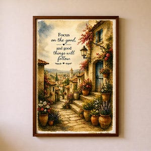 Op de afbeelding: Een ingelijste kunstdruk met een mediterrane dorpsscène in aquarel-stijl met stenen trappen, terracotta potten en bloeiende bloemen. De tekst luidt: Focus on the good, and good things will follow. De lijst is van donkerbruin hout.