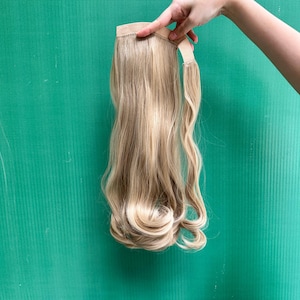 May include: A long, wavy synthetic hair ponytail extension in a light blonde shade with darker root tones. The hairpiece features a wrap-around attachment strip at the top, designed for securing the extension to natural hair with a smooth finish.