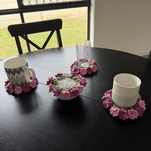 May include: Three round crochet coasters featuring a floral design with pink, purple, and green yarn petals. One coaster supports a patterned mug, another holds a clear ribbed glass, and the third sits under a plain white mug on a black table.