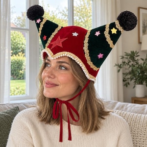 May include: A two-pointed velvet hat in red and dark green featuring black pom-poms at the tips, gold braided trim, white pearl bead edging, and small star-shaped appliqués. The hat includes thin red velvet ties that fasten under the chin.