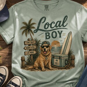 May include: A sage green t-shirt featuring a graphic of a golden retriever wearing sunglasses and a baseball cap. The design includes a palm tree, a surfboard, a cooler, and wooden signs that read Stay Salty, Work Less, and Live More.