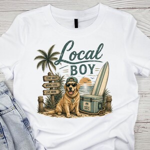 May include: A white t-shirt featuring a graphic of a golden retriever wearing a baseball cap and sunglasses. The design includes a palm tree, a surfboard, a cooler, and a wooden sign with the text Local Boy, Stay Salty, Work Less, Live More.