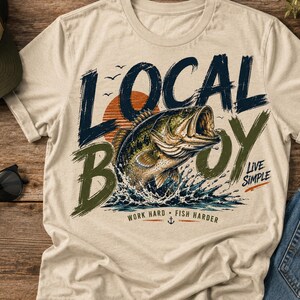 May include: A beige t-shirt featuring a graphic of a largemouth bass jumping out of water. The text reads LOCAL BOY, LIVE SIMPLE, and WORK HARD FISH HARDER. The design includes a sunset, birds, and an anchor icon in dark blue and olive green.
