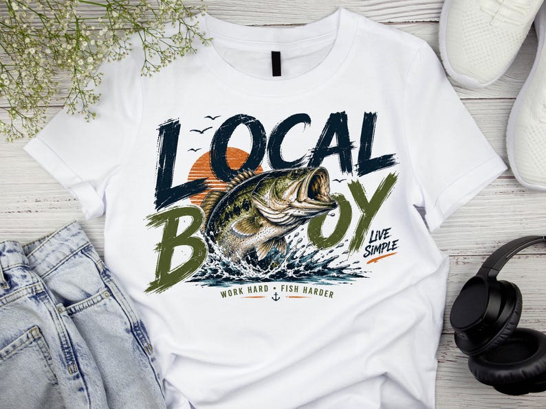 May include: A white t-shirt featuring a graphic of a jumping bass fish in front of an orange sun. The text reads LOCAL BOY, LIVE SIMPLE, and WORK HARD FISH HARDER with a small anchor icon. The design uses dark blue, olive green, and orange colours.