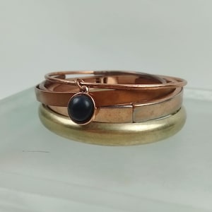 May include: A stack of three bangles featuring a smooth gold-tone band, a flat copper-tone band, and a thin copper-tone wire band. A small, round, black cabochon charm hangs from the thin wire band, adding a dark contrast to the metallic tones.